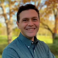 Smiling man in a checkered shirt standing outdoors with trees in the background.