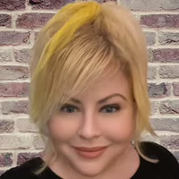 Professional headshot of a woman with blonde hair styled in a casual look, smiling against a textured brick background.