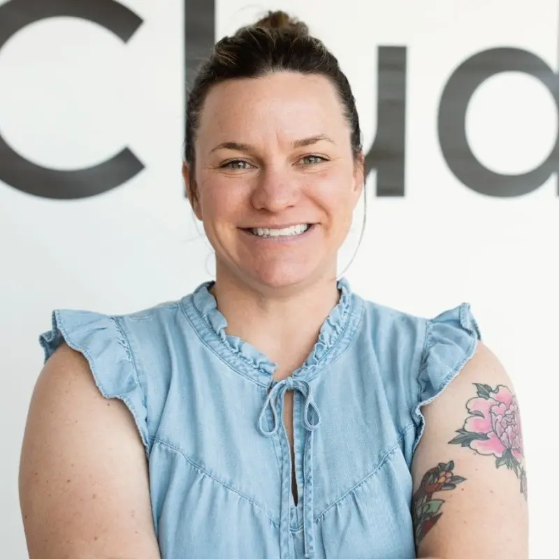 A smiling woman with a floral tattoo on her arm, wearing a denim top, poses in front of a large "Club" sign.