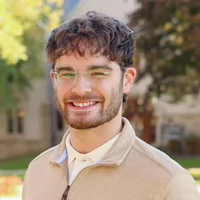 A smiling young man with curly hair wearing glasses and a beige sweater, standing outdoors with a blurred background of trees and buildings.