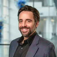 Headshot of a smiling man with a beard, wearing a black shirt and gray blazer, set against a blurred background.