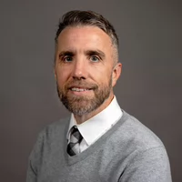 Professional headshot of a man with a beard, wearing a gray sweater and a white shirt with a checked tie.