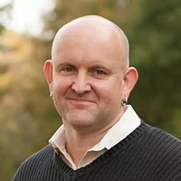 Professional portrait of a man with a shaved head and earrings, wearing a black sweater and smiling outdoors.