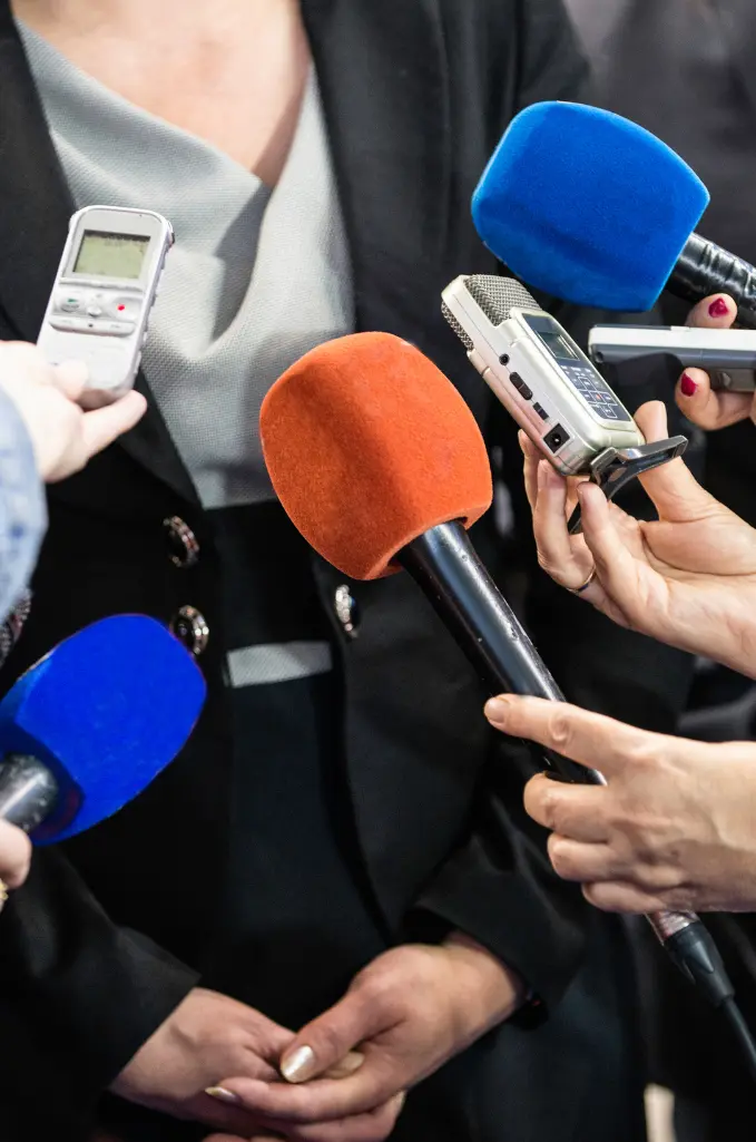 Microphones of various colors surround a person, indicating a press conference or media event.