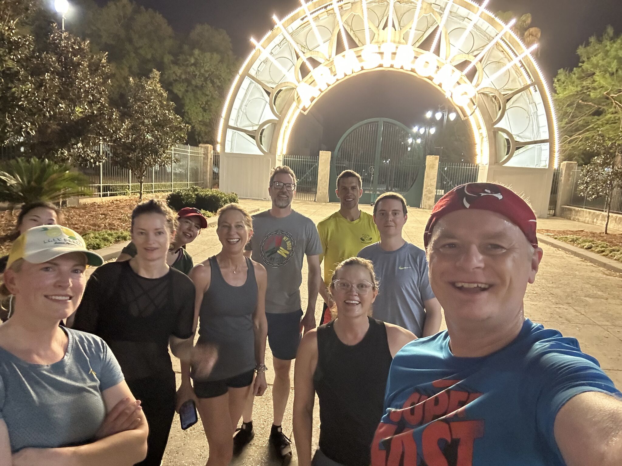 Group selfie of runners in front of the Armstrong Park entrance at night, featuring bright lights and a lively atmosphere.