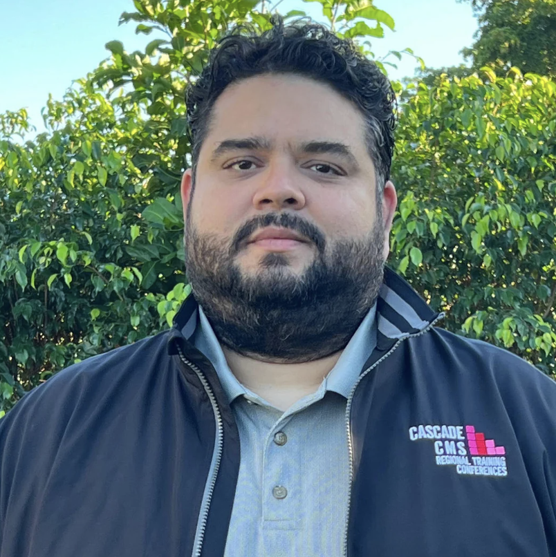 A man with a beard wearing a dark jacket with "Cascade CMS" logo stands outdoors against a backdrop of greenery.